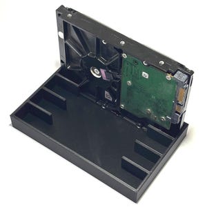 May include: A black plastic storage tray with internal dividers, holding a single rectangular hard drive disk in an upright position. The hard drive features a green circuit board, metallic casing, and visible connection ports on the side.