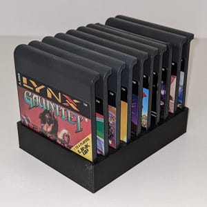 Puede incluir: Un soporte de cartuchos de juego de plástico negro con varios cartuchos de juego Atari Lynx. El cartucho frontal muestra el juego "Gauntlet" con un gráfico de guerrero. El soporte está diseñado para exhibir y almacenar los cartuchos de juego.