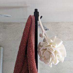 May include: A white bath pouf hanging on a black rack with a red towel. The pouf is made of soft, fluffy material and is designed for use in the shower or bath.