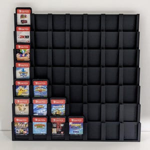 May include: Black wall-mounted storage rack with 48 slots for video game cartridges. The rack is filled with various Nintendo Switch game cartridges, including titles like "2K18", "Sonic Colours", and "WWE 2K18".