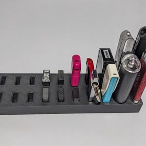 Puede incluir: Un organizador rectangular negro con múltiples ranuras, que contiene varios artículos como encendedores, llaves y pequeños dispositivos electrónicos. Los artículos son de diferentes colores y formas, creando una solución de almacenamiento funcional.