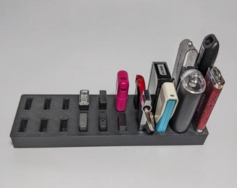 May include: A black, rectangular organizer with multiple slots, holding various items like lighters, keys, and small electronic devices. The items are in different colors and shapes, creating a functional storage solution.