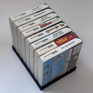 May include: A selection of Nintendo DS game cartridges in clear cases, held in a black stand. Titles visible include "Cooking Guide", "Zoo Hospital", "Nintendogs", "Madagascar: Escape 2 Africa", and "New Super Mario Bros."