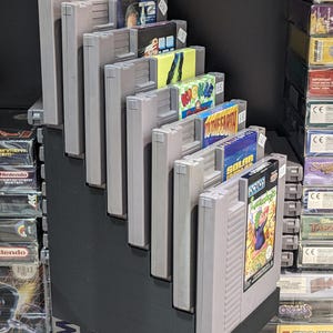 Puede incluir: Una exhibición de cartuchos de juegos de Nintendo Entertainment System (NES) vintage. Los cartuchos son grises con etiquetas de juegos coloridas, dispuestos en un estante negro escalonado. Algunas etiquetas son visibles, incluyendo "Solar" y "To The Earth".