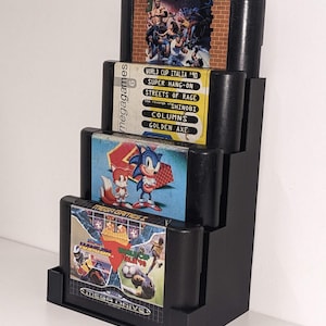 May include: A black stepped display stand holding four Sega Mega Drive game cartridges. The cartridges feature game titles including "World Cup Italia '90", "Sonic the Hedgehog", and "Streets of Rage". The stand is designed to showcase the games.