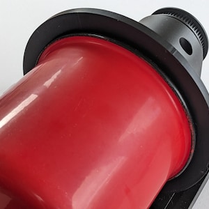 May include: A close-up of a red metal can with a black plastic ring around the top. The can is being held in place by a black plastic device.