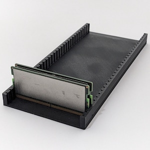 Könnte beinhalten: Ein schwarzer 3D-gedruckter RAM-Halter mit zwei eingesetzten RAM-Riegeln. Der Halter hat eine rechteckige Basis mit vertikalen Stützen an den Seiten und einem strukturierten Innenraum. Die RAM-Riegel haben grüne Leiterplatten und silberne Metallkomponenten.