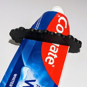 Può includere: Spremi-tubo per dentifricio in plastica nera con un design ondulato. Lo spremi-tubo è su un tubo di dentifricio Colgate blu e rosso.