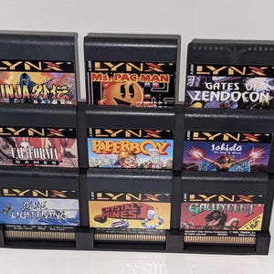 May include: A black plastic game cartridge holder filled with nine Atari Lynx game cartridges. The cartridges feature colorful game titles like "Ninja Gaiden," "Ms. Pac-Man," and "Gates of Zendocon." The holder is designed to display the game cartridges.