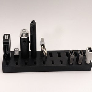 May include: Black USB charging station with multiple slots for charging various devices. The station has a white, black, and silver USB drive, a black flashlight, and a black memory card reader.