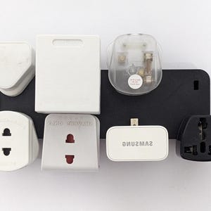 May include: A collection of various white and black travel adapter plugs, including a Samsung-branded adapter. The adapters are arranged on a black rectangular base against a white background. The plugs are designed for international use.