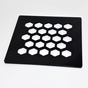 Op de afbeelding: Een zwart, vierkant object met een honingraatpatroon van zeshoekige uitsparingen. Het object is gemaakt van een solide, ondoorzichtig materiaal en staat tegen een witte achtergrond. De uitsparingen zijn gelijkmatig verdeeld, waardoor een rasterachtig ontwerp ontstaat.