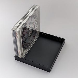 Könnte beinhalten: Ein schwarzes CD-Aufbewahrungsetui mit einer Reihe von Trennwänden. Das Etui enthält mehrere CDs in transparenten Kunststoffhüllen. Die CDs haben schwarz-weiße Kunstwerke. Das Etui steht auf einer weißen Oberfläche.