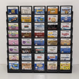 May include: A black display stand filled with Nintendo DS game cartridges. The cartridges feature various game titles, including "High School Musical," "Brain Training," and "Hello Kitty." The stand is organised in rows and columns, showcasing a variety of game covers.