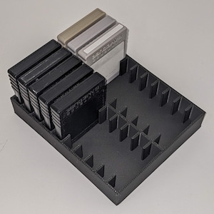 May include: A black 3D-printed game cartridge organizer holding several black, gray, and beige Nintendo GameCube game cartridges. The organizer has multiple slots for storage.