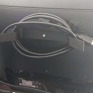 May include: Black cable tidy mounted on the back of a computer monitor. The tidy is a black bracket with a screw, holding a coiled black cable with an HDMI connector.