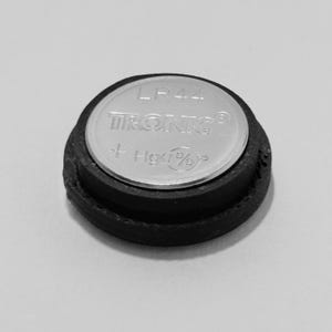 Op de afbeelding: Een ronde, zilverkleurige LR44 Tronic knoopcelbatterij met een zwarte rubberen ring als basis. Het bovenoppervlak bevat de tekst LR44, TRONIC, een plusteken en Hg 0%, gedrukt in een helder, in reliëf aangebracht metallic lettertype.