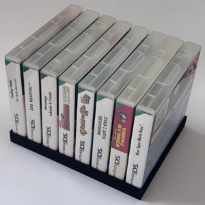 Könnte beinhalten: Ein schwarzer, rechteckiger Spielhalter mit sieben Nintendo DS-Spielhüllen. Die Spielhüllen haben transparente Kunststoffabdeckungen und verschiedene Spieltitel, darunter "Cooking Guide", "Zoo Hospital" und "Kung Fu Panda".