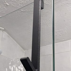 May include: A black metal shower door hook. The hook is designed to hang over a glass shower door. The hook has a simple, functional design with a textured finish.