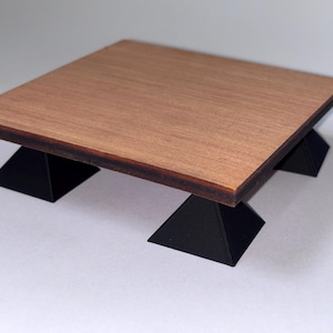 May include: A small, square wooden table with a light brown top and dark brown edges. The table is supported by four black, triangular legs. The table is approximately 10 inches square and 4 inches tall.