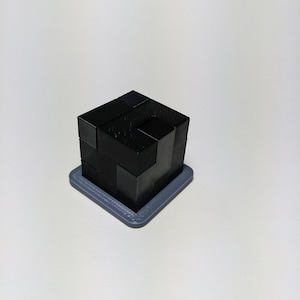 May include: A black 3D-printed puzzle cube with interlocking geometric shapes, resting on a grey square base. The puzzle is designed for assembly and disassembly, a brain teaser.