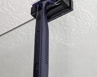 May include: A black razor with a blue and clear plastic head is mounted on a glass surface with a black plastic holder.