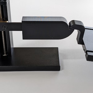 Könnte beinhalten: Ein schwarzes Metallgerät mit rechteckiger Basis, vertikaler Stütze mit Feder und verstellbarem Arm. Der Arm ist mit einem Scharnier verbunden. Das Gerät scheint ein Präzisionswerkzeug oder -instrument zu sein.