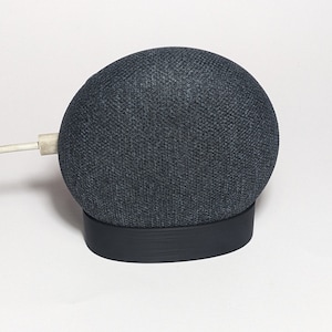 May include: A dark gray fabric-covered smart speaker with a black base. The speaker is plugged in with a white cord.