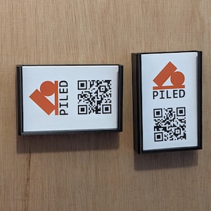 May include: Two rectangular black framed signs with the word "PILED" in orange and a QR code. The signs also feature an orange abstract design. The signs are on a light brown wooden surface.