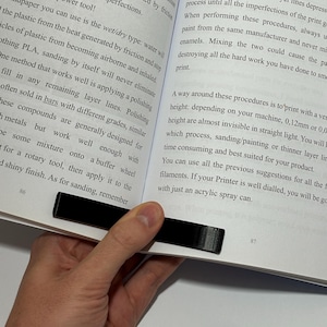 May include: A black, rectangular, 3D-printed plastic book page holder clip positioned at the bottom centre of an open book. The clip features a smooth, solid finish and is designed to hold the pages of the book open while reading.