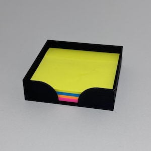 May include: A black square desk tidy containing a pad of neon yellow sticky notes. The organiser has curved cutouts at the front and a visible stack of colourful sticky notes. The background is a plain, light grey.