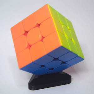 Könnte beinhalten: Ein 3x3-Würfelpuzzle mit orangefarbenen, gelben und blauen Seiten. Der Würfel steht auf einem schwarzen Ständer.