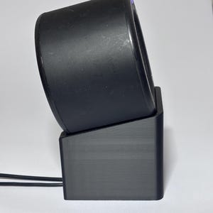 Könnte beinhalten: Ein schwarzes, zylindrisches Gerät mit abgerundeter Oberseite, das auf einem schwarzen, rechteckigen Sockel ruht. Das Gerät ist leicht angewinkelt, wodurch ein Blick ins Innere möglich ist. Zwei schwarze Drähte gehen von der Basis aus.