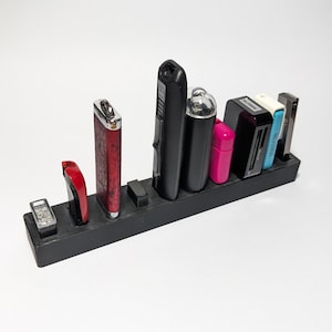 Puede incluir: Un organizador rectangular negro que contiene varios encendedores y pequeños dispositivos electrónicos. Los encendedores son rojos, negros y plateados. Otros artículos incluyen una funda rosa y un lector de tarjetas negro. El organizador es de material negro.