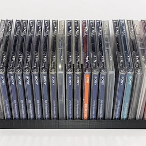 Puede incluir: Un estante de almacenamiento de plástico negro lleno de estuches de DVD transparentes. Los estuches tienen etiquetas azules y blancas con el logotipo "SONY". Algunos estuches tienen etiquetas naranjas con "TDK" impreso. Los estuches están apilados ordenadamente.