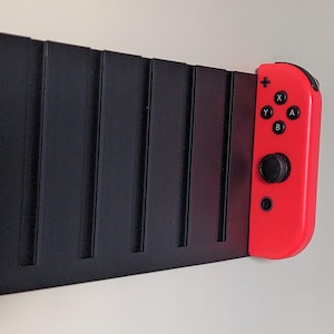 May include: A black wall mount with a red Nintendo Switch Joy-Con controller attached. The controller has the standard buttons: X, Y, A, B, and a directional pad.