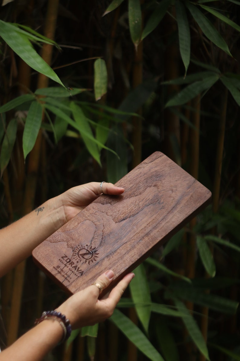 K&ouml;nnte beinhalten: Ein Schneidebrett aus Holz mit einer nat&uuml;rlichen Holzmaserung. Das Brett ist mit dem Text "Zdrava" und einem Logo mit einer Sonne und einem Buchstaben Z graviert. Das Brett wird von den H&auml;nden einer Person gehalten.
