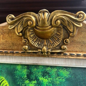May include: A decorative gold-toned frame featuring an ornate, raised central crest with symmetrical scrollwork and a sunburst pattern. The frame has a textured, light-coloured inner border with a dark dotted line detail along the edge.
