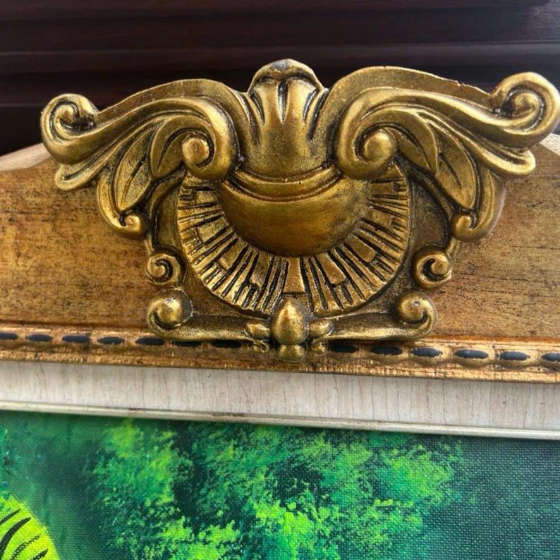 May include: A decorative gold-toned frame featuring an ornate, raised central crest with symmetrical scrollwork and a sunburst pattern. The frame has a textured, light-coloured inner border with a dark dotted line detail along the edge.
