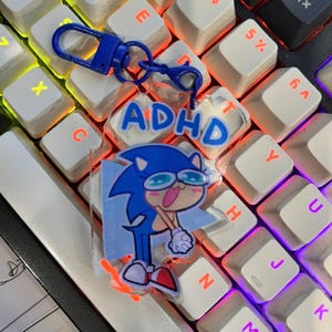 Pode incluir: Um chaveiro azul com uma imagem de desenho animado de Sonic the Hedgehog usando óculos e a palavra "ADHD". O chaveiro está preso a um fecho azul e está em um teclado branco.