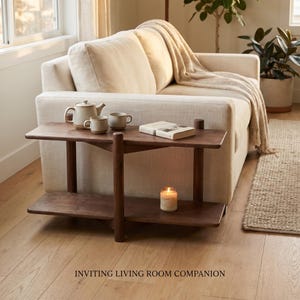 May include: A two-tiered dark wood console table featuring a rectangular top and bottom shelf. The top surface holds a ceramic teapot, two matching mugs, and a closed book. The bottom shelf displays a single lit candle in a glass jar.