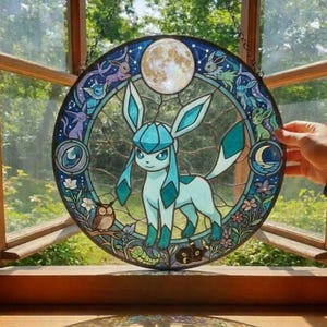 Może przedstawiać: Okrągłe witrażowe okno przedstawiające postać Glaceona w odcieniach niebieskiego i turkusowego, otoczone innymi postaciami Pokémonów, pełnią księżyca i elementami kwiatowymi. Okno wisi przed oknem.