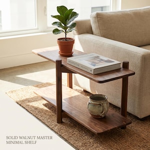 May include: A two-tier solid walnut shelf with rounded edges and four cylindrical legs. The top tier holds a potted green plant and a book titled Architecture. The bottom tier holds a ceramic pot. Text reads: SOLID WALNUT MASTER MINIMAL SHELF.