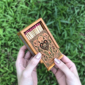 May include: A wooden matchbox with a floral design and the text "The Perfect Match" and "M & A 2024". The matchbox is filled with matches with red tips.