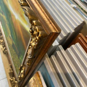 May include: An ornate gold-coloured picture frame with intricate scrollwork and relief detailing. The frame surrounds a painting depicting a landscape with green hills and a bright yellow sunburst. The frame has a metallic finish and angled edges.