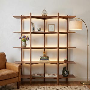 May include: A modern, wooden bookshelf with built-in lighting. The shelves are filled with books, vases, and decorative items. A leather armchair and a floor lamp with a beige shade are positioned nearby, creating a cozy reading nook.