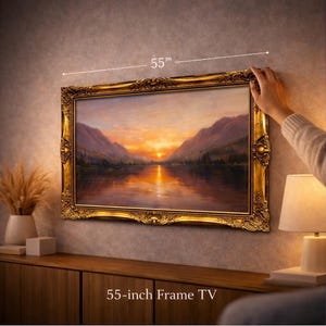 May include: A 55-inch frame TV featuring an ornate, metallic gold-coloured frame with intricate scrollwork and floral detailing. The screen displays a landscape painting of a sunset over a calm lake surrounded by mountains in shades of orange.