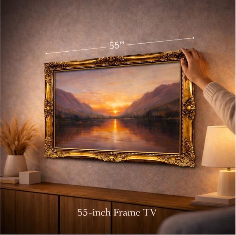 May include: A 55-inch frame TV featuring an ornate, metallic gold-coloured frame with intricate scrollwork and floral detailing. The screen displays a landscape painting of a sunset over a calm lake surrounded by mountains in shades of orange.