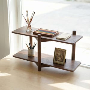 May include: A two-tiered, dark wood shelf unit with various art supplies. The top shelf holds a glass jar with paint brushes, a wooden tray of colored pencils, and a small notebook. The bottom shelf displays a framed photo and a ceramic cup with pencils.