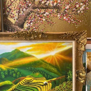 May include: A framed oil painting depicting a vibrant sunrise over green terraced rice fields and mountains, set within an ornate gold frame. Above it, a partial view of another painting shows a bird perched on a flowering tree branch.